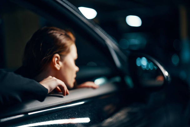 een nauwe horizontale foto van de zijkant, 's nachts, van een vrouw die in een zwarte auto zit en uit het raam kijkt en in de zijspiegel kijkt. Hoge kwaliteit foto - Foto, afbeelding