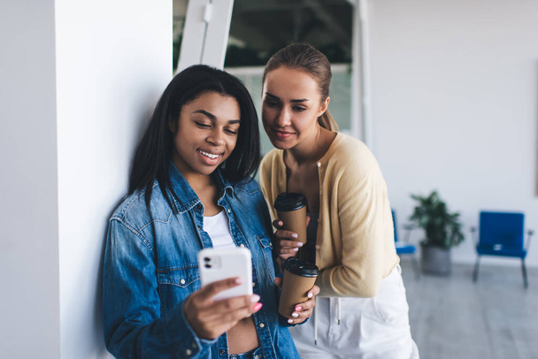 Multiracial businesswomen watching something on mobile phone during work in office. Concept of modern successful women. Idea of teamwork. Young smiling european and black girls with coffee - Zdjęcie, obraz