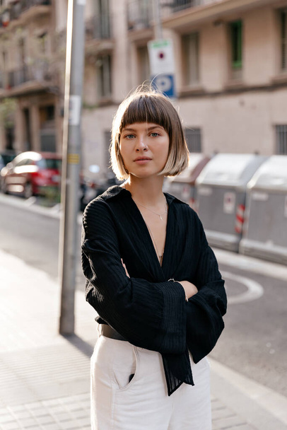 Outside portrait of stylish pretty European girl with straight short hairstyle wearing black blouse and white pants posing at camera in sunlight on summer street - Foto, Imagen