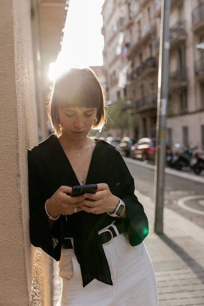 incredible charming woman with short straight hairstyle wearing black blouse using smartphone and enjoying walking in the city in summer warm sunny evening.  - Photo, image