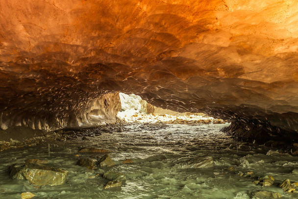 Grotta di ghiaccio arancione scavata nel ghiacciaio Vallelunga attraversata da un torrente. Il ghiacciaio è in rapido ritiro a causa del riscaldamento globale, Alto Adige, Italia - Foto, immagini