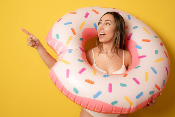 Portrait of an excited brunette young woman dressed in swimsuit holding float ring pointing finger side isolated over yellow background. - Foto, Imagen