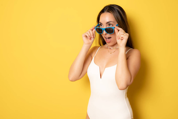 Young happy excited brunette woman in swimsuit wearing sunglasses looking side standing isolated over yellow background. Summer holidays concept. - 写真・画像