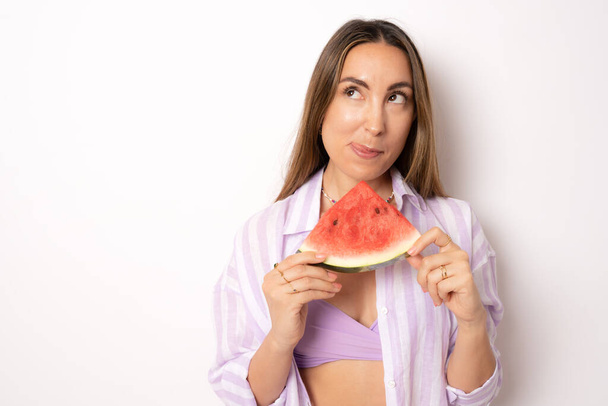 bikini Woman leaning against white background holding watermelon up to mouth - Фото, изображение