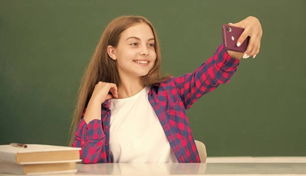 happy kid making selfie on smartphone in classroom at blackboard, portrait. - Photo, image