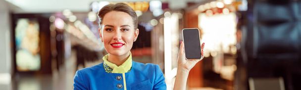 Vreugdevolle vrouw stewardess in lucht gastvrouw uniform kijken naar de camera en glimlachen terwijl het houden van moderne smartphone - Foto, afbeelding