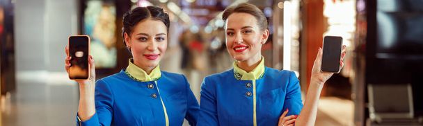 Fröhliche Flugbegleiterinnen in Stewardess-Uniform blicken in die Kamera und lächeln, während sie ihr Handy in der Hand halten - Foto, Bild