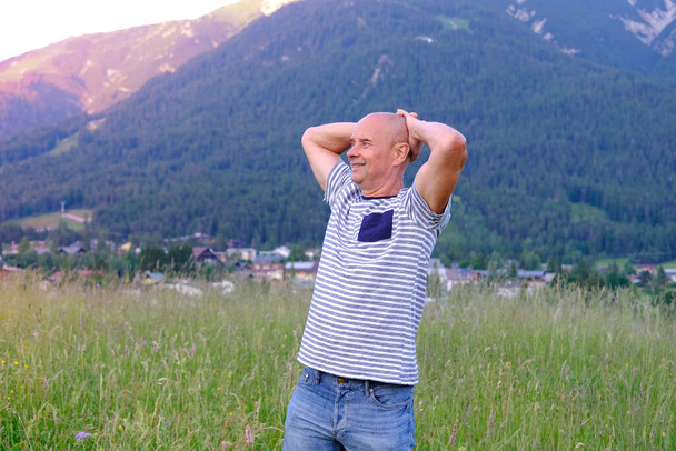 mature man, senior 60 years old standing in mountains above city, smiles, Alps are behind him, green grass in meadow, concept of enjoy life in old age, travel, active lifestyle, human happiness - Photo, image
