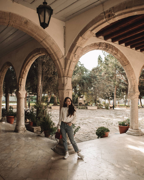 A young woman is standing near the beautiful building with arch, lanterns and potted plants in Panagia Chrisopolitissa, Larnaca, Cyprus.  - Foto, imagen