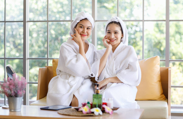 Zwei junge, schöne asiatische Kurgäste in weißem, sauberem Bademantel und Handtuch sitzen lächelnd zusammen und berühren gesunde Wangen und Gesicht auf dem Sofa nach einer Aromatherapie-Massage-Sitzung. - Foto, Bild