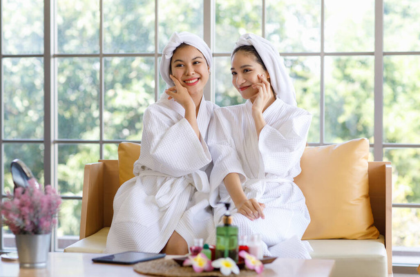 Zwei junge, schöne asiatische Kurgäste in weißem, sauberem Bademantel und Handtuch sitzen lächelnd zusammen und berühren gesunde Wangen und Gesicht auf dem Sofa nach einer Aromatherapie-Massage-Sitzung. - Foto, Bild
