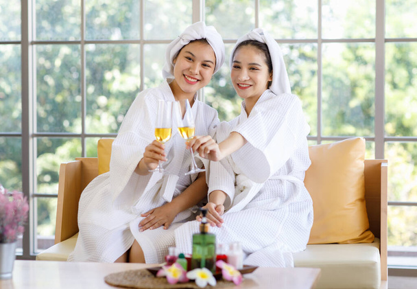 Two Asian young beautiful spa customers friend in white clean bathrobe covered head with towel sitting smiling holding drinking fruit juice beverage glasses talking together after therapy session. - Photo, Image