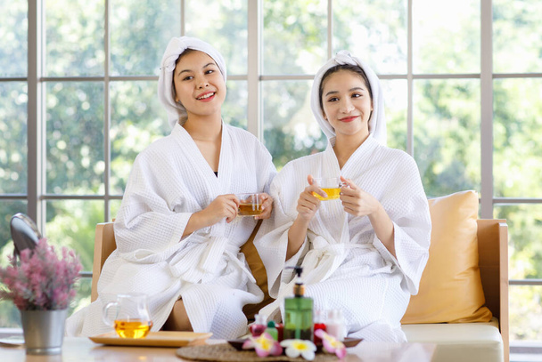 Two Asian young beautiful spa customers in white clean bathrobe covered head with towel sitting smiling holding cheers toasting hot herbal tea beverage glasses drinking together after therapy session. - Photo, Image