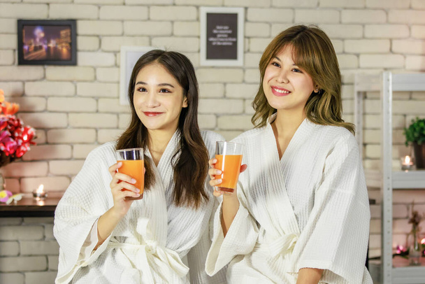 Two Asian young beauty happy female customers in white clean massaging bathrobe towel sitting resting relaxing smiling hold orange juice glasses cheers together on massage bed before therapy session. - Photo, Image