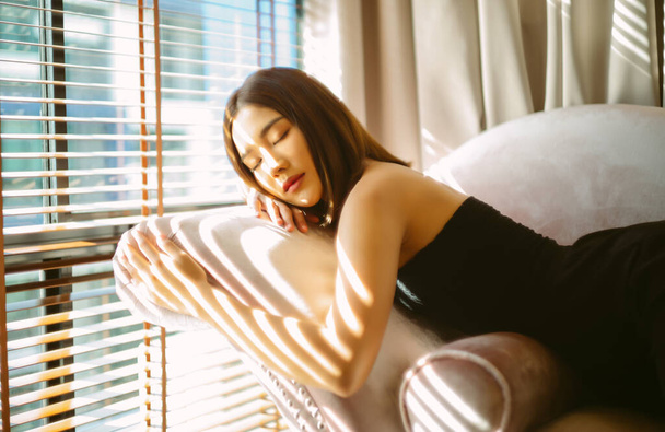 Beautiful sexy Asian woman in stylish dress shoulder clothes and closed eyes while lying on sofa in living room with sunlight on her face. Fashion look, beauty and style. Cozy domestic atmosphere. - Fotó, kép