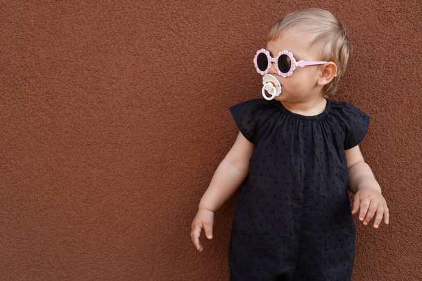Cute little girl wearing stylish clothes with sunglasses and pacifier near brown wall. Space for text - Foto, Bild
