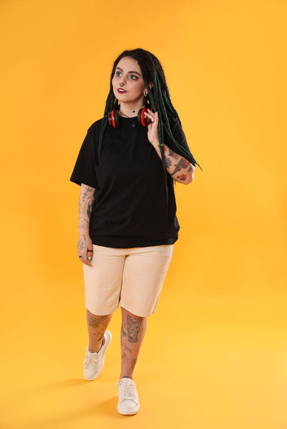 Beautiful young woman with tattoos on body, nose piercing and dreadlocks against yellow background - Zdjęcie, obraz