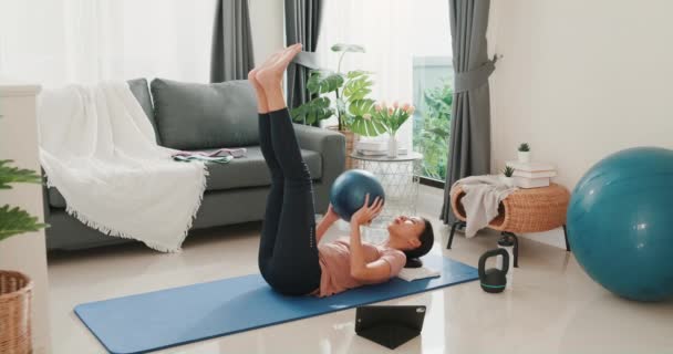 Young attractive Asia woman in sportswear watching fitness online video on tablet with fitness ball in living room at house. Home quarantine workout and fitness exercise without the gym. - Imágenes, Vídeo