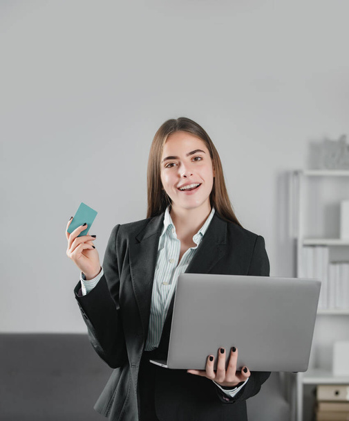 Financial services, credit card banking concept. Manager holding laptop computer and credit card indoors in office. Young business woman financial planning - Fotó, kép