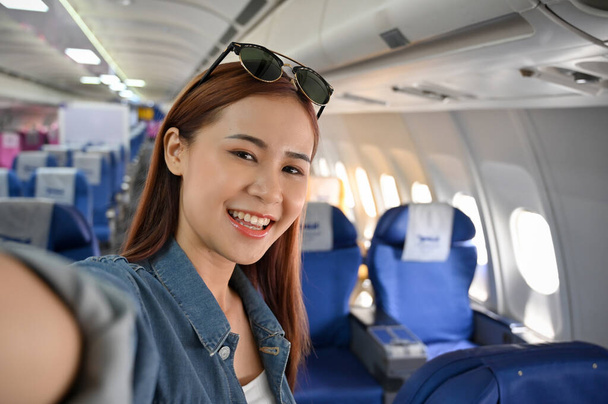 Μια όμορφη ευτυχισμένη νεαρή ασιάτισσα ταξιδιώτισσα ή τουριστική σέλφι στο αεροπλάνο κατά τη διάρκεια ενός ταξιδιού σε κάποιο μέρος. Ταξίδι, ταξίδι, κορίτσι στο πλοίο έννοια. - Φωτογραφία, εικόνα