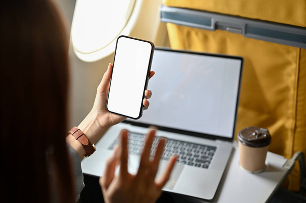 Close-up image, Female passenger at the window seat in economy class using portable laptop and smartphone during the flight. Phone white screen mockup. - Photo, Image