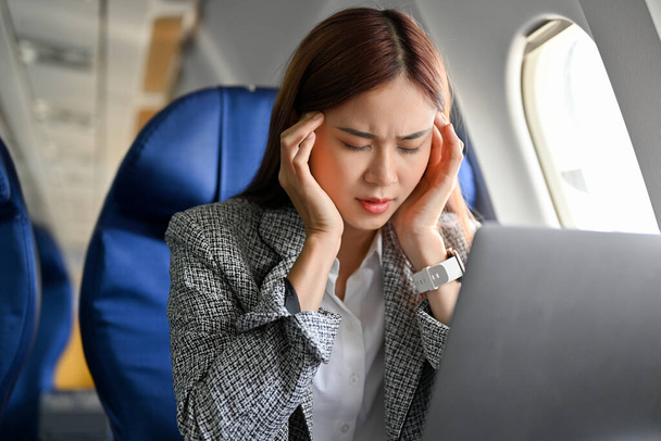 Stressato e grave giovane donna d'affari asiatica a distanza che lavora sull'aereo, soffre di mal di testa e mal di aria durante il volo. - Foto, immagini