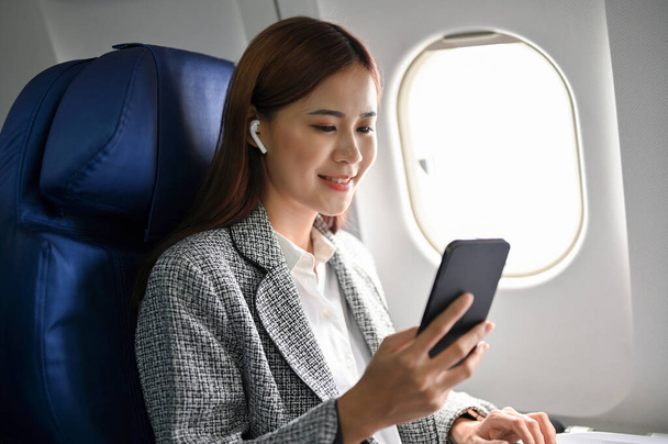 Piękna azjatycka bizneswoman siedzi na swoim miejscu w samolocie za pomocą smartfona, aby przeczytać wiadomości biznesowe lub e-mail podczas lotu. Koncepcja transportu lotniczego i biznesmenów. - Zdjęcie, obraz
