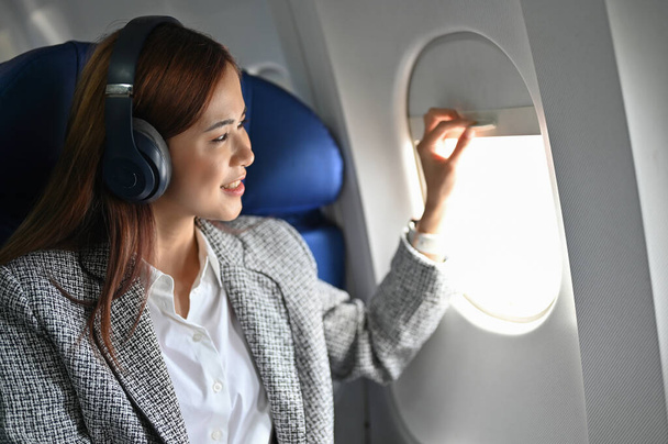 Hermosa y atractiva joven mujer de negocios asiática o mujer con auriculares inalámbricos se sienta en el asiento de la ventana levantando una ventana de avión para mirar una hermosa vista del cielo. - Foto, imagen