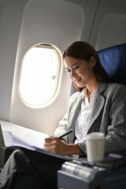 Nádherná a profesionální asijská podnikatelka nebo výkonná manažerka ve formálním obleku sedí na svém sedadle obchodní třídy a čte si zprávu o návrhu schůzky, přičemž si během letu zapisuje do dokumentu. - Fotografie, Obrázek