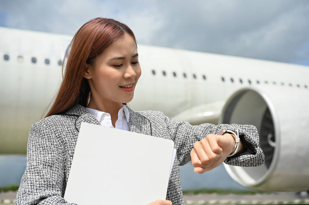 Eine schöne junge asiatische Sekretärin oder Assistentin, die die Uhrzeit an der Armbanduhr überprüft, vor dem Flugzeug steht und auf ihren Chef oder Geschäftskunden wartet. Geschäftsflug oder Geschäftsreisekonzept - Foto, Bild