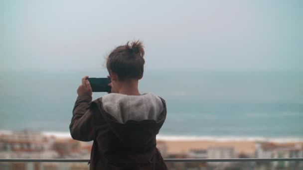 Niño viajero tomando fotos con teléfono inteligente desde el balcón. Escena oceánica y playa con hoteles en Nazare town resort, Portugal. Disparo con enfoque cambiante - Metraje, vídeo