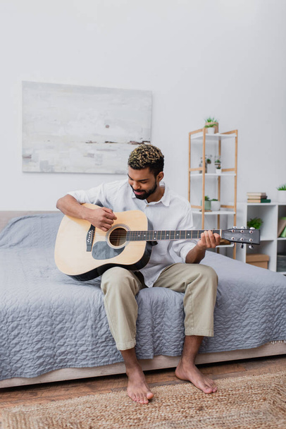 happy young african american man with dyed hair and beard playing acoustic guitar in bedroom - Foto, Imagem