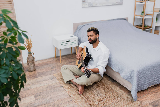 vousatý africký Američan s obarvenými vlasy sedí na koberci a hraje na akustickou kytaru u postele - Fotografie, Obrázek