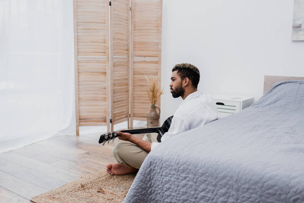 бородатий афроамериканський чоловік сидить на килимі і грає на акустичній гітарі біля ліжка
 - Фото, зображення