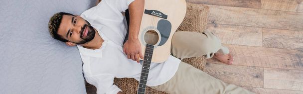 horní pohled na potěšeného afrického Američana s obarvenými vlasy držící akustickou kytaru a opírající se o postel, banner - Fotografie, Obrázek