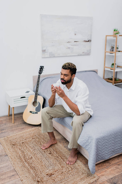 high angle view of african american man holding cup of coffee while sitting on bed near acoustic guitar - Photo, image