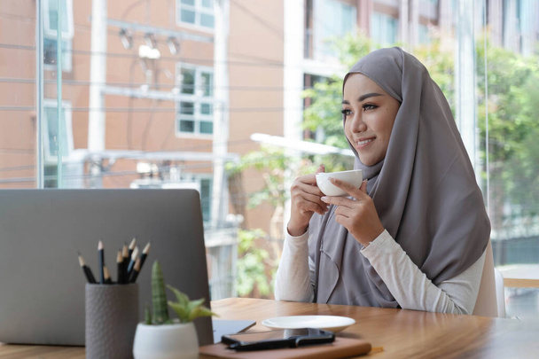 lächelnd schöne muslimische Geschäftsfrau, die heißen Kaffee trinkt, im Arbeitsbereich sitzt und an Fenster außerhalb denkt, Arbeitsplan denkt - Foto, Bild