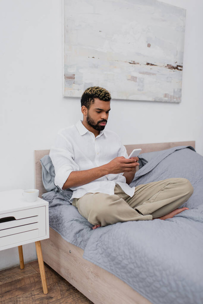 молодой африканский американец с окрашенными волосами с помощью смартфона во время отдыха на кровати - Фото, изображение