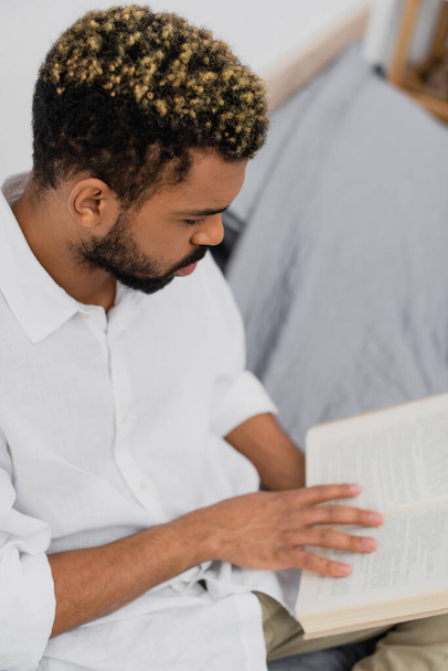 vista de ángulo alto de barbudo hombre afroamericano con teñido libro de lectura de pelo en el dormitorio - Foto, Imagen