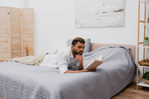 młody Afroamerykanin człowiek z farbowanych włosów czytania książki leżąc na łóżku - Zdjęcie, obraz