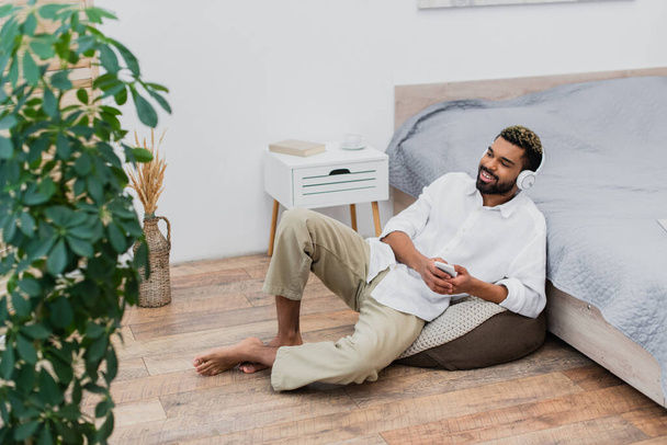 felice uomo africano americano in cuffie wireless ascoltare musica mentre tiene smartphone e seduto vicino al letto - Foto, immagini