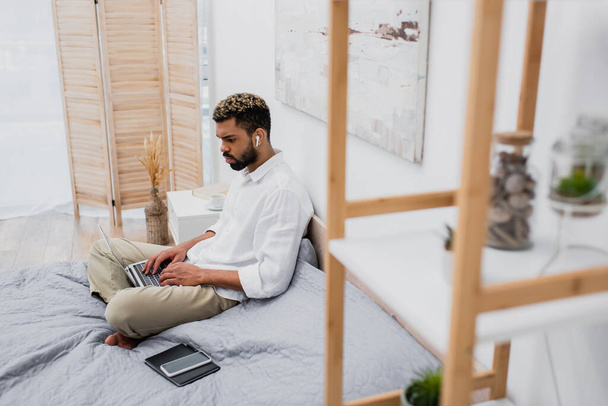 giovane uomo africano americano in auricolare senza fili utilizzando il computer portatile mentre seduto sul letto a casa - Foto, immagini