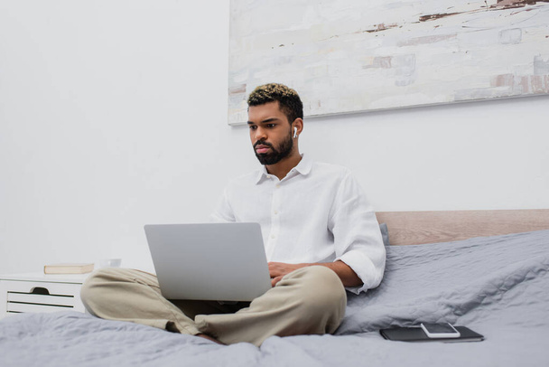 young african american freelancer in wireless earphone using laptop while sitting on bed at home - Photo, Image