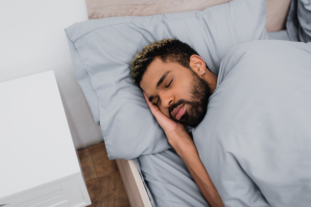 Yüksek açılı, sakallı, gözleri kapalı, yatağın yanında uyuyan Afro-Amerikan bir adam. - Fotoğraf, Görsel