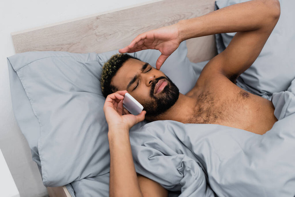 high angle view of awakened african american man talking on smartphone in bed - Photo, Image