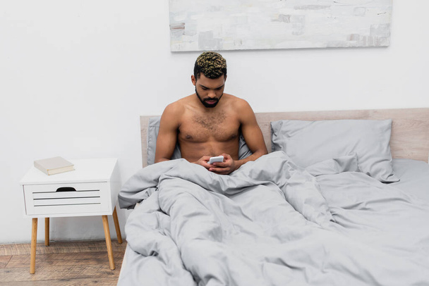 shirtless african american man with dyed hair chatting on smartphone in bedroom  - Fotografie, Obrázek