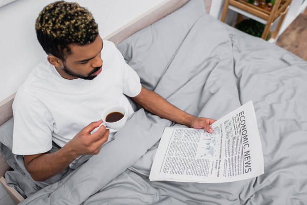vista de ángulo alto del hombre afroamericano con el pelo teñido sosteniendo la taza de café mientras lee el periódico en la cama - Foto, Imagen