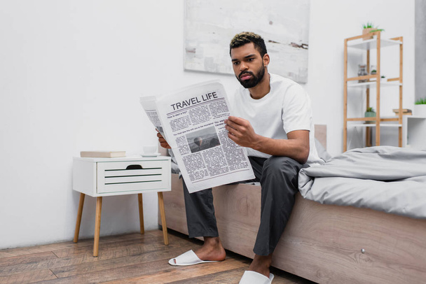 african american man with dyed hair sitting on bed and reading travel life newspaper - Фото, изображение
