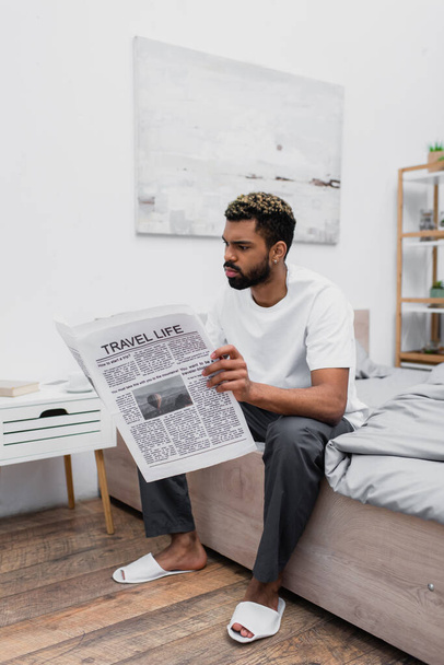 sério afro-americano homem com cabelos tingidos sentado na cama e ler viagem vida jornal - Foto, Imagem