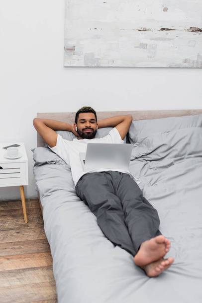 homem americano africano alegre em fones de ouvido sem fio assistindo filme enquanto deitado na cama com laptop  - Foto, Imagem